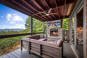 A covered outdoor seating area with a stone fireplace, a television mounted on the wall, and a view of the landscape. - Fire on the Mountain - Mountain Views | 3 Levels of Decks | Outdoor Fireplaces | Hot Tub | Game Room (Mineral Bluff)