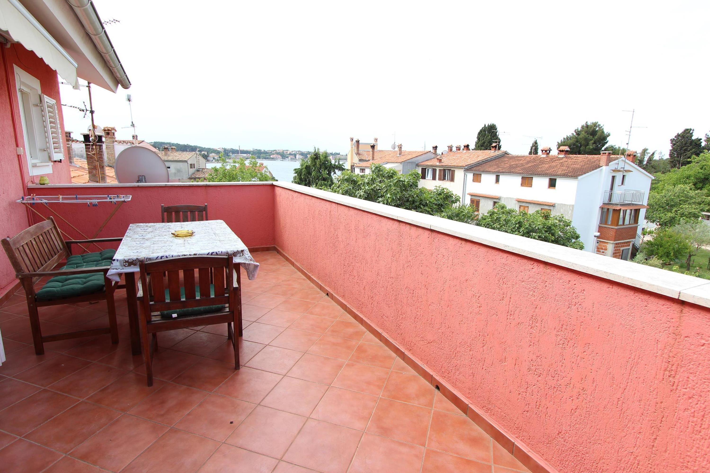 A balcony with a table and chairs, a red wall, and a view of buildings and trees.
