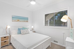 A bedroom with a bed, two bedside lamps, a nightstand, a wall-mounted painting, a window with blinds, and a ceiling fan.