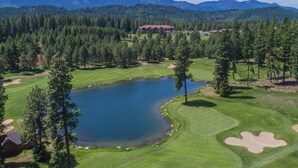 Campo de golf con un estanque, rodeado de pinos y montañas.