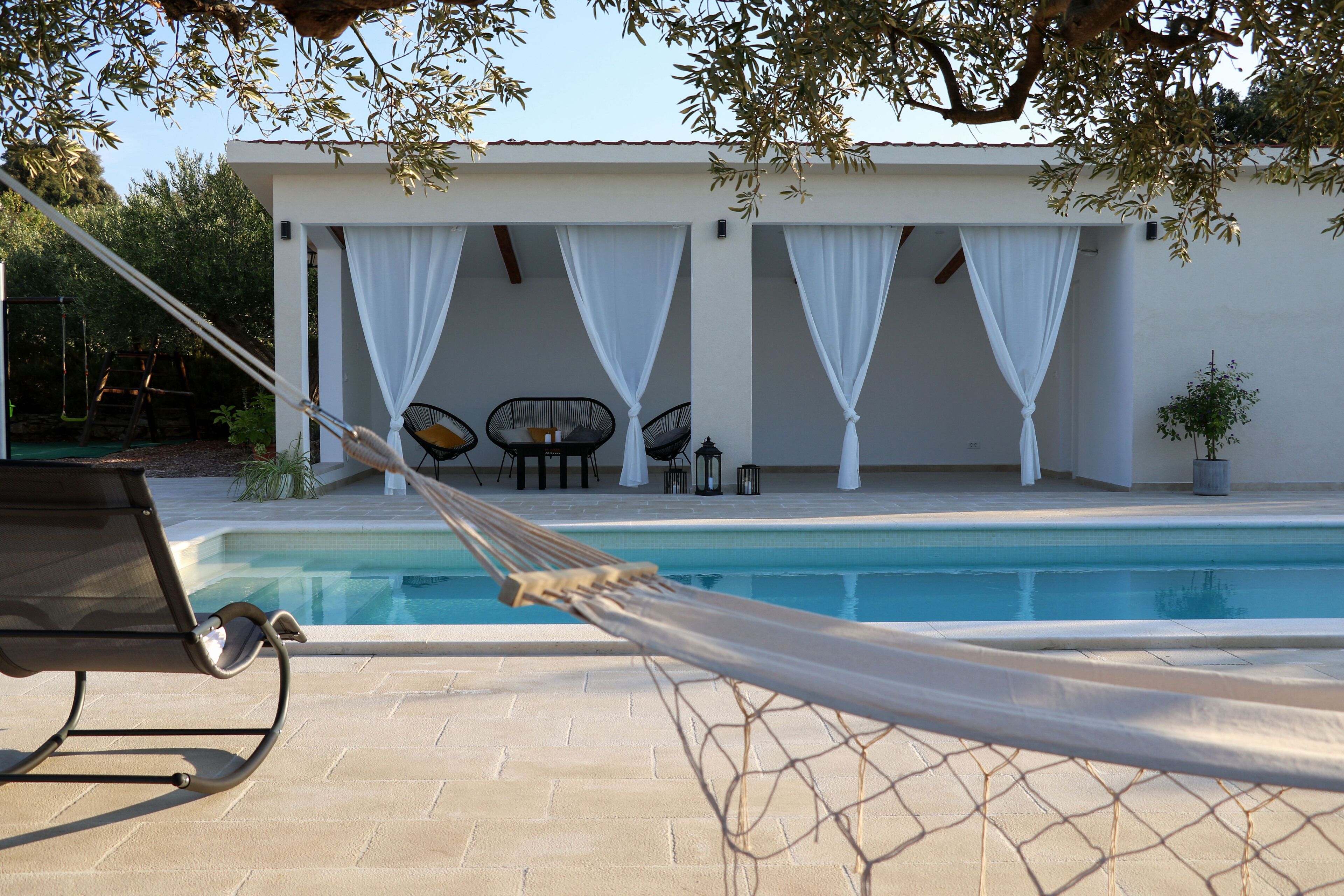 A poolside area with a hammock, lounge chairs, and a covered seating area with white curtains.