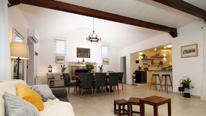 Amplia sala de estar con mesa de comedor, sillas, sofá con cojines, una araña y una cocina al fondo.
