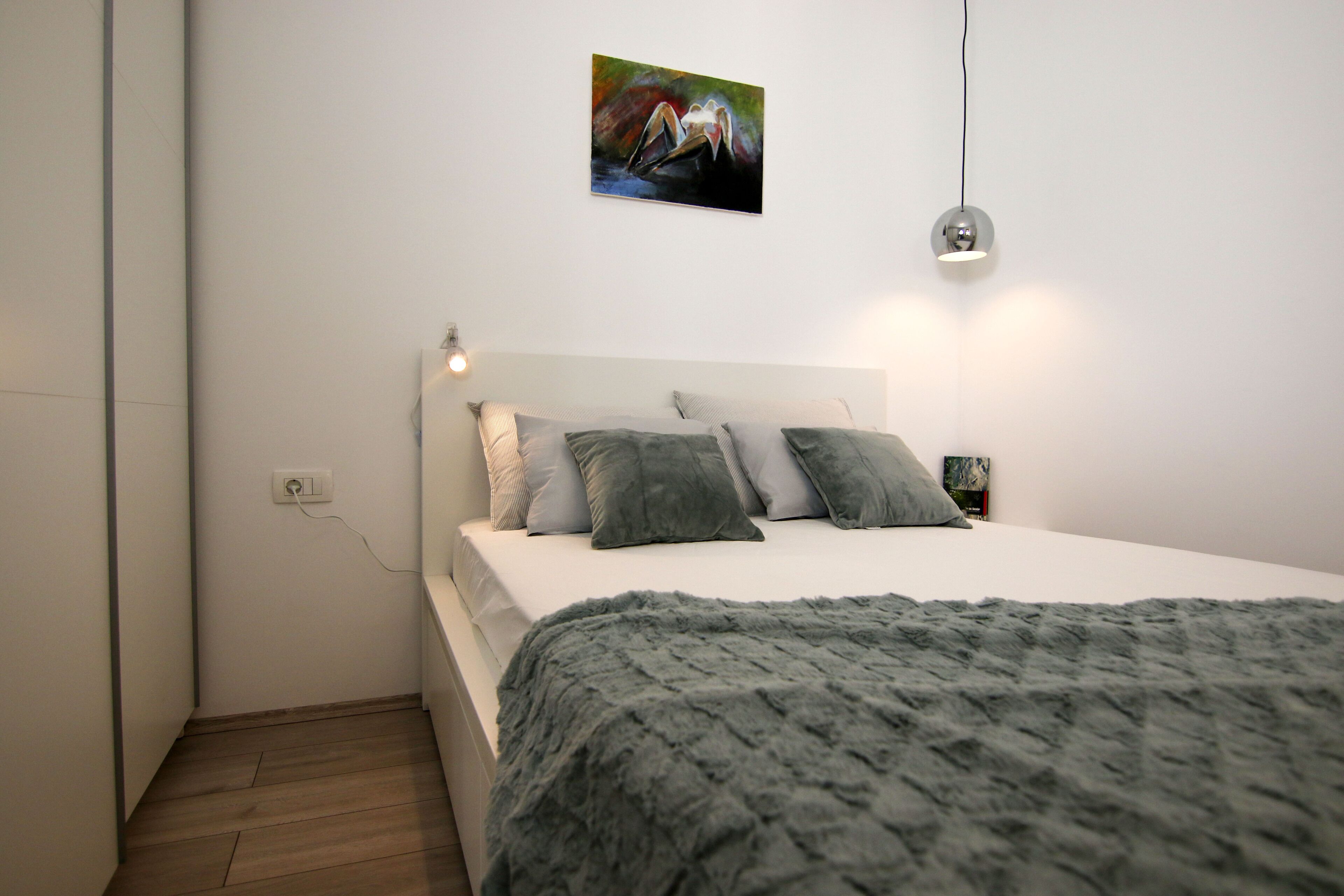A modern bedroom with a white bed, grey pillows, a grey blanket, a painting on the wall, and a bedside lamp.