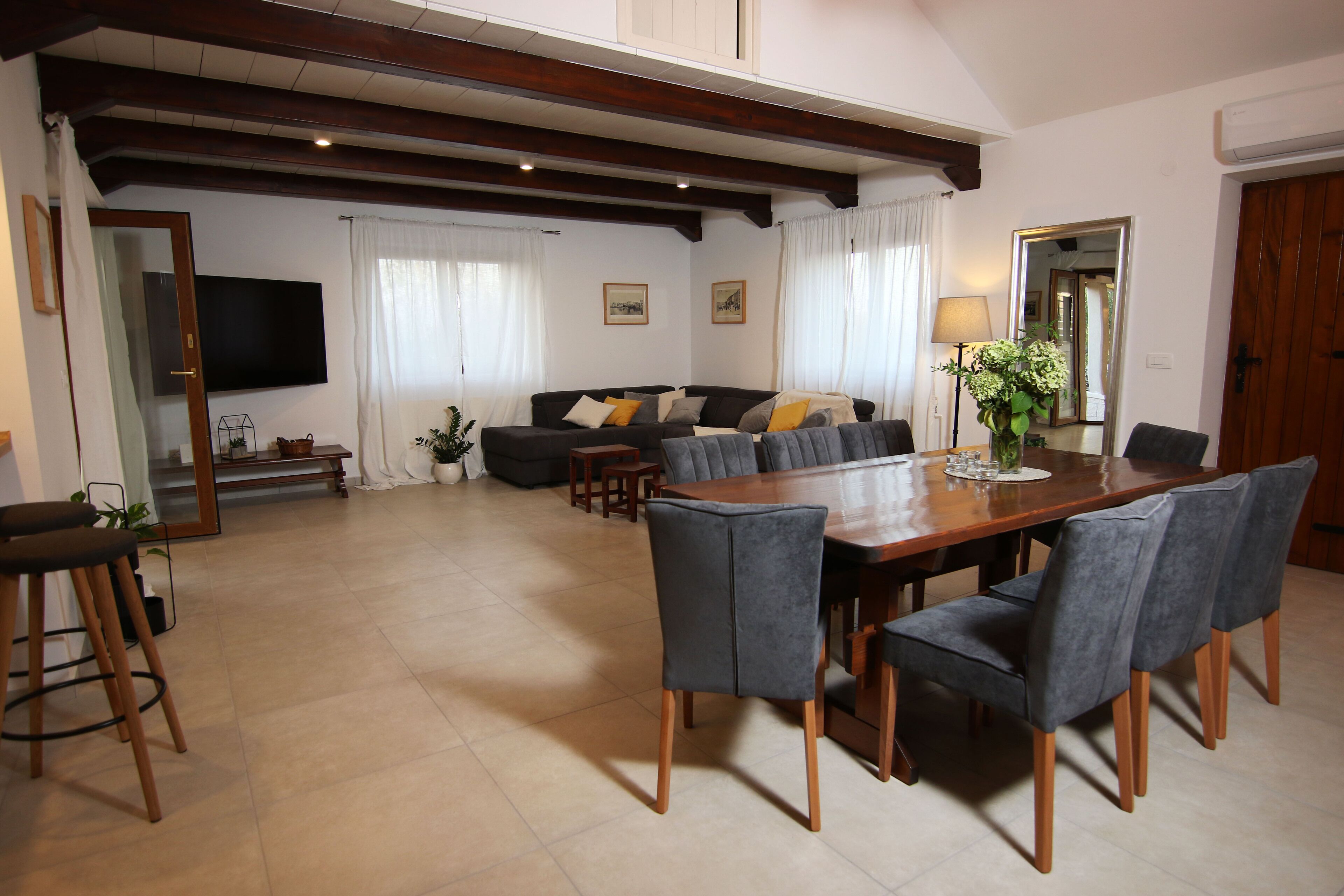 Un vaste salon comprenant une table à manger en bois, des chaises à revêtement gris, un canapé avec des coussins, un téléviseur à écran plat fixé au mur et un grand miroir qui reflète la pièce.