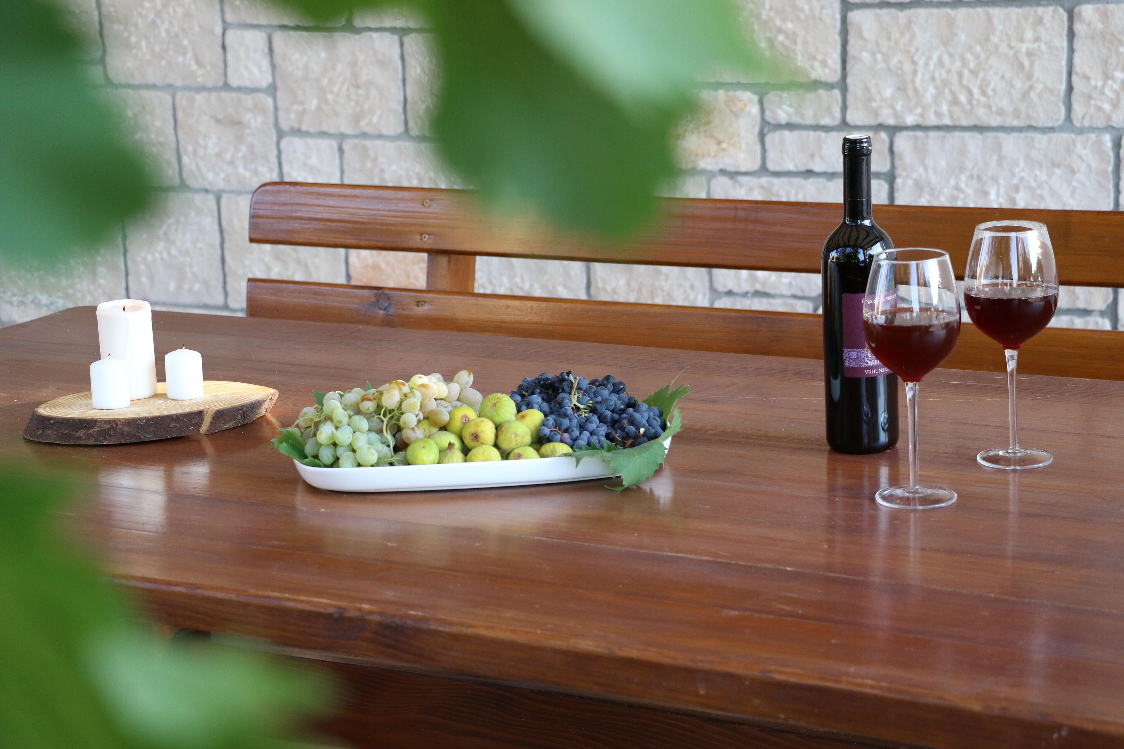 A wooden table with a bottle of wine, two glasses of red wine, and a plate of grapes and other fruits.