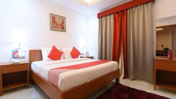A hotel room with a bed, two bedside tables, a red throw, a red rug, a painting on the wall, and a window with curtains.