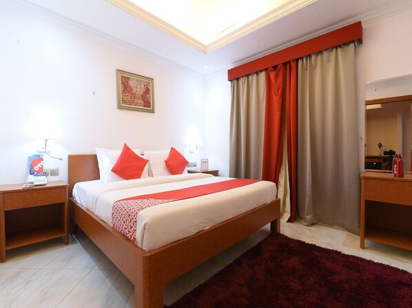 A hotel room with a bed, two bedside tables, a red throw, a red rug, a painting on the wall, and a window with curtains.