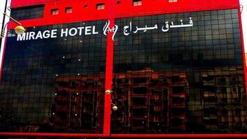 Un edificio moderno con una amplia fachada de vidrio y la inscripción 'MIRAGE HOTEL' en inglés y árabe.