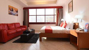 Ein Hotelzimmer mit einem roten Sofa, einem Bett mit rot-weißer Bettwäsche, einem Nachttisch mit Lampe und einem Fenster mit Vorhängen.