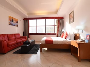 Quarto de hotel com sofá vermelho, cama com roupa de cama em vermelho e branco, mesa de cabeceira com abajur e janela com cortinas.