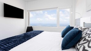 Ein modernes Hotelzimmer mit einem großen Bett, einem flachbildfernseher an der Wand und einem großen Fenster mit Blick auf den Himmel und die Wolken.