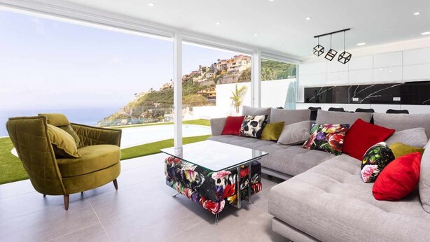 Ein modernes Wohnzimmer mit Sofa, Sessel und Couchtisch, große Fenster mit Blick auf eine Hügellandschaft und eine minimalistische Küche.