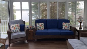 Een knus ingerichte kamer met een groot raam met uitzicht op een meer, voorzien van rieten meubels en kleurrijke kussens.