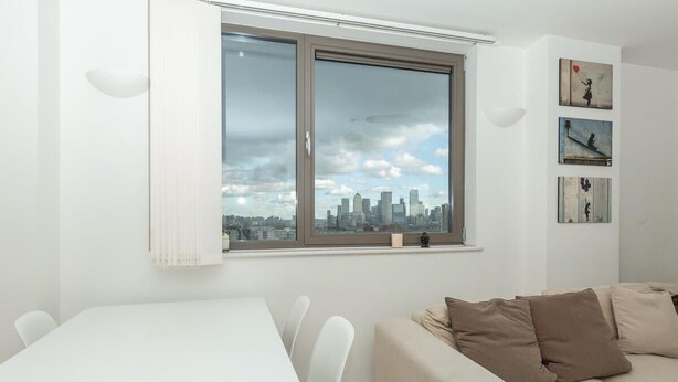 A modern living room with a dining area, a large window with a city view, and a wall with three framed pictures. - Stunning East London 2 Bed Apartment With Views (London)
