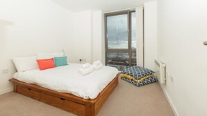 A bedroom with a wooden bed, white bedding, and a view through a large window. - Stunning East London 2 Bed Apartment With Views (London)