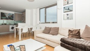 A modern living room with a sofa, a coffee table, and a dining area with chairs. There is a kitchen in the background with a washing machine and cabinets. - Stunning East London 2 Bed Apartment With Views (London)