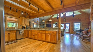 A spacious kitchen with wooden cabinets and a large island, a dining area with a table and chairs, and a view of the outdoors through large windows. - Spectacular Views, Privacy and Hot Tub (Maggie Valley)