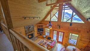 A cozy cabin interior with a stone fireplace, wooden ceiling beams, a dining area, and large windows offering mountain views. - Spectacular Views, Privacy and Hot Tub (Maggie Valley)
