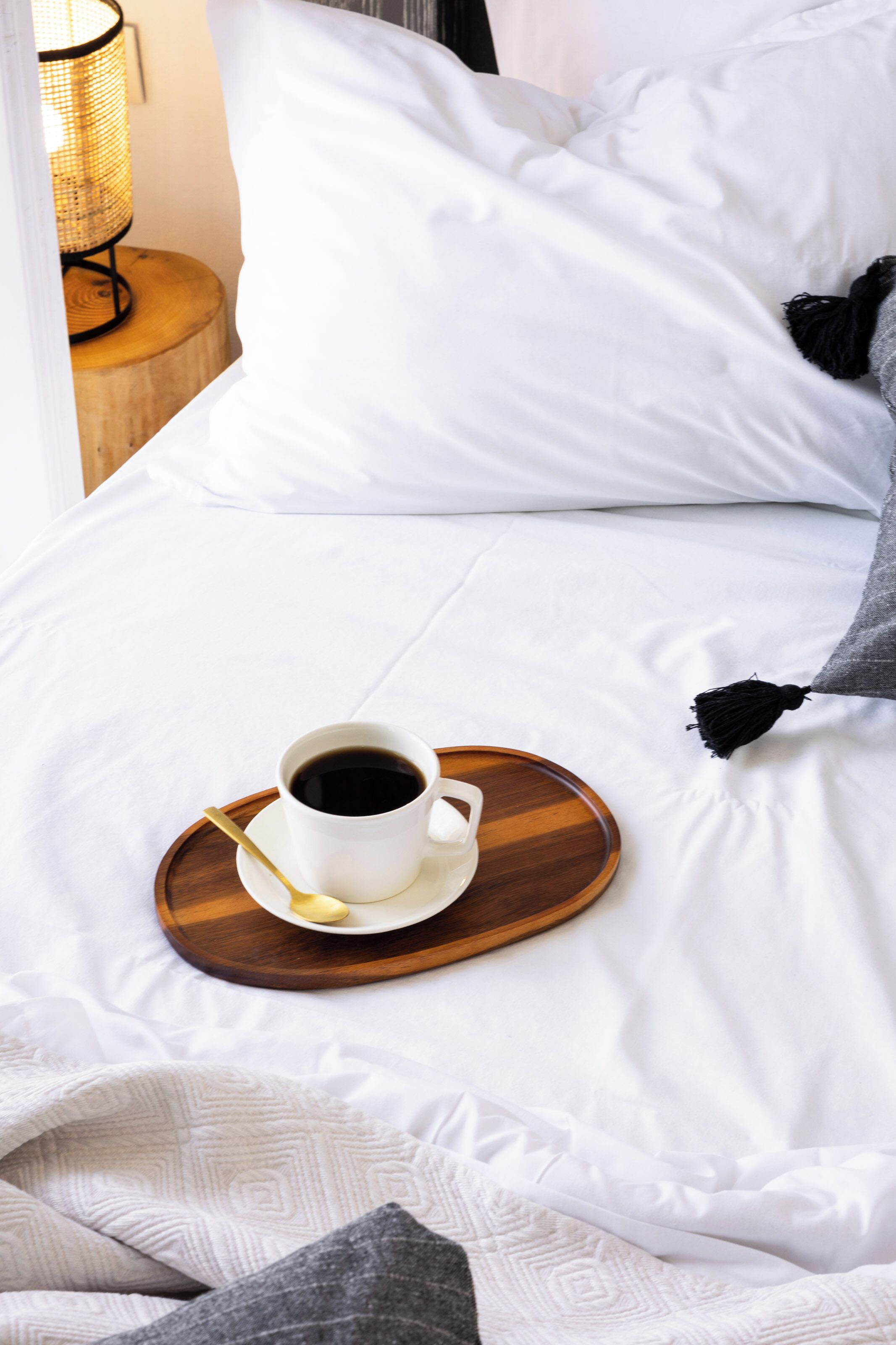 A neatly made bed with white linens, a cup of coffee on a wooden tray, and a bedside lamp.