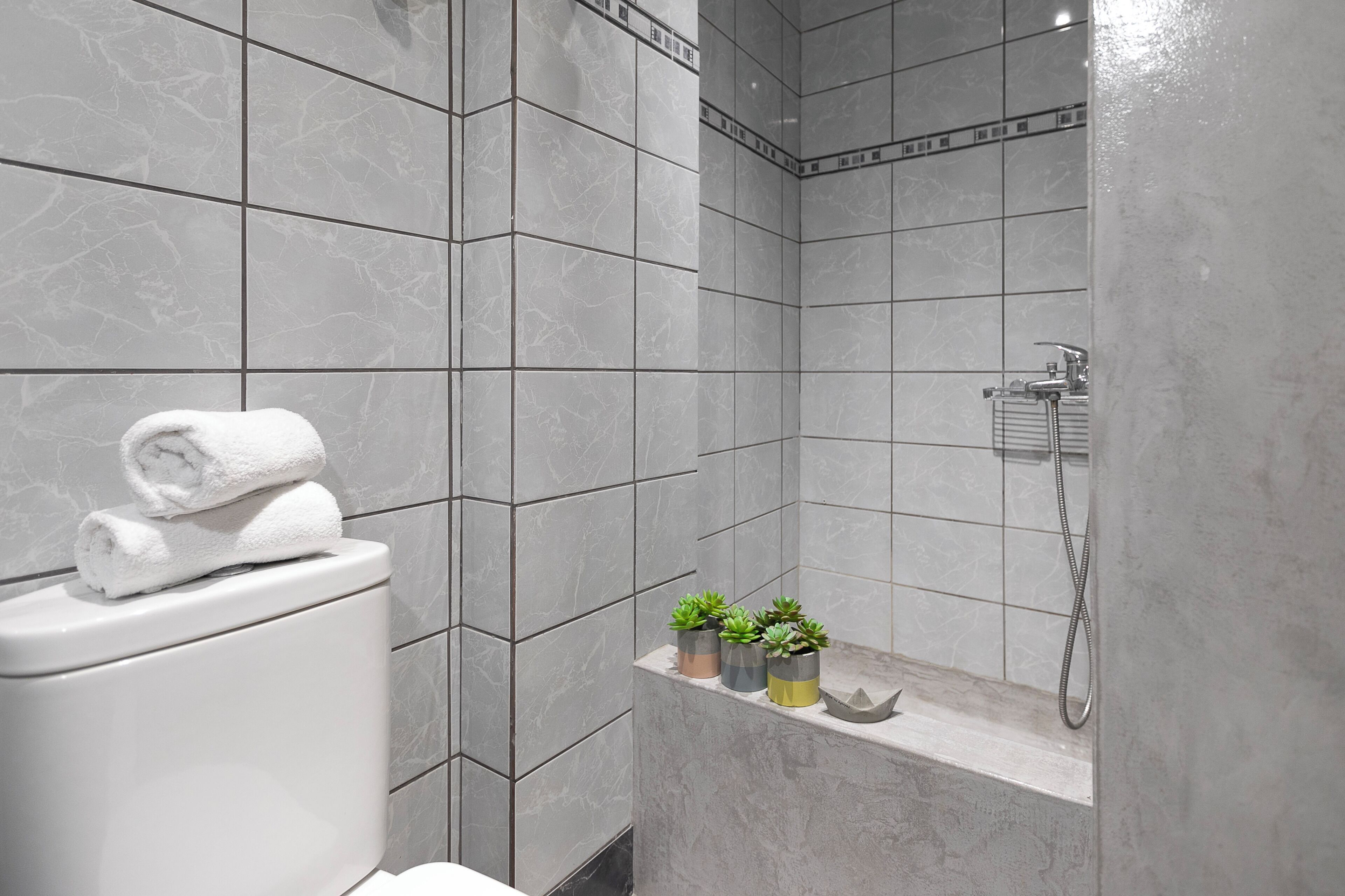 Une salle de bain moderne avec des carreaux gris, des toilettes avec une serviette roulée, une douche et trois petites plantes en pot disposées sur une étagère.