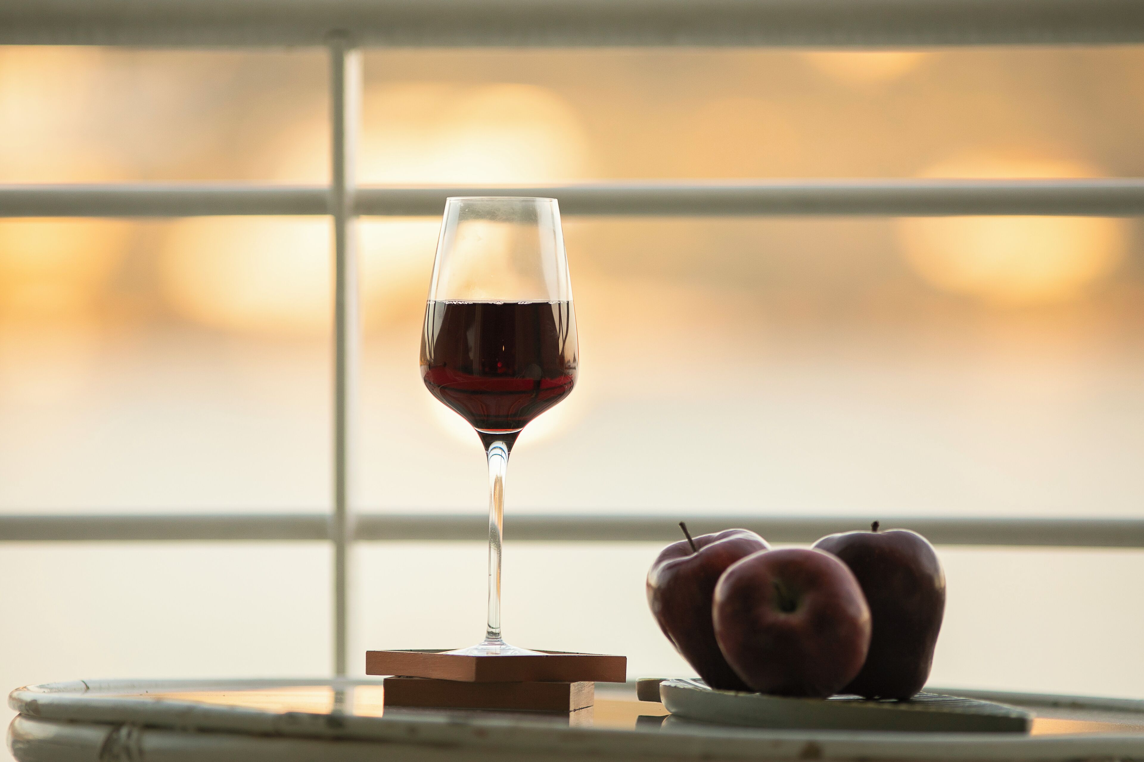 Una copa de vino sobre un posavasos junto a dos manzanas sobre una mesa cerca de una ventana.
