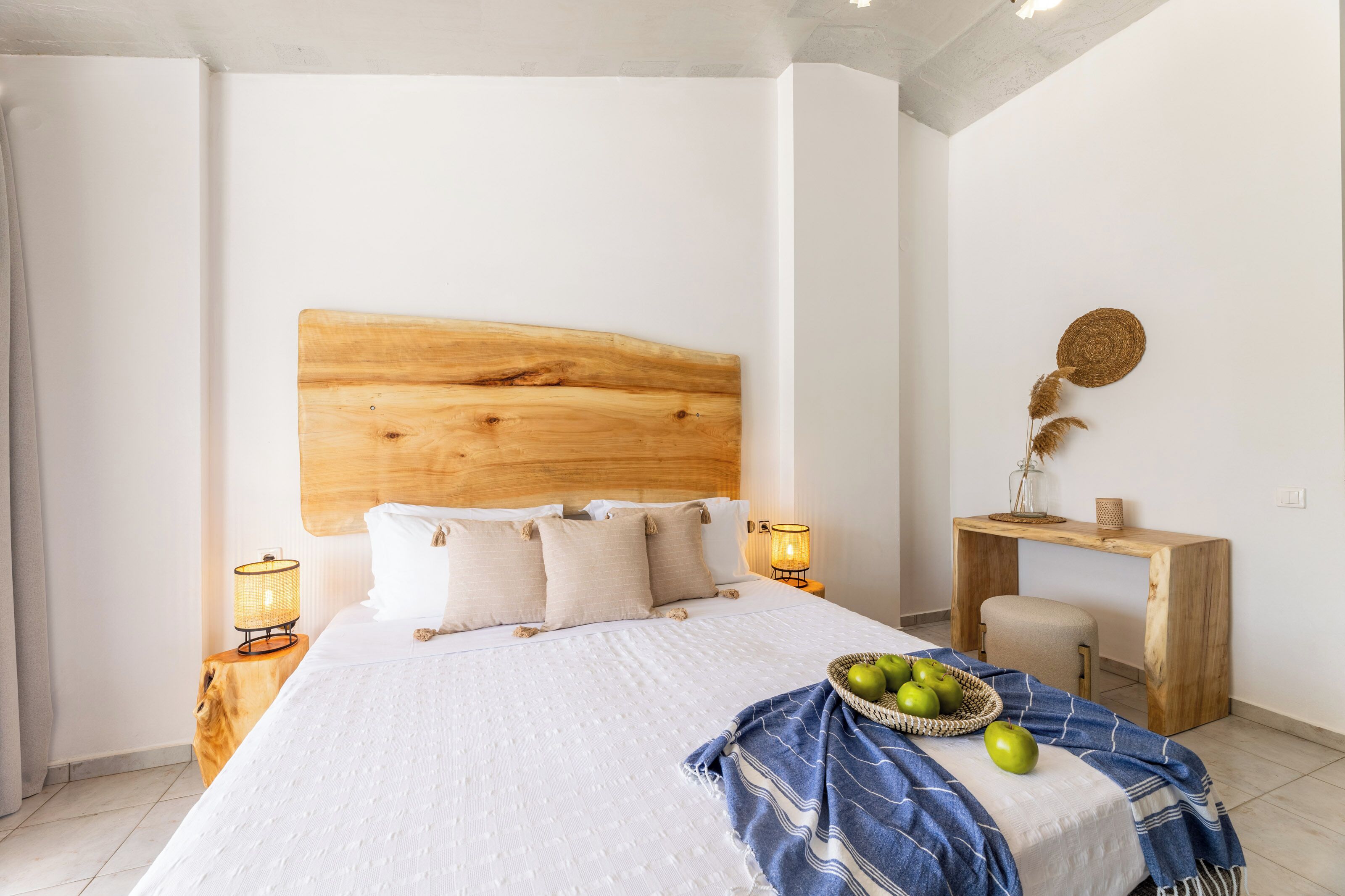 A bedroom with a wooden headboard, white bedding, a bedside table with a lamp, a small desk with a chair, and a basket of apples on the bed.