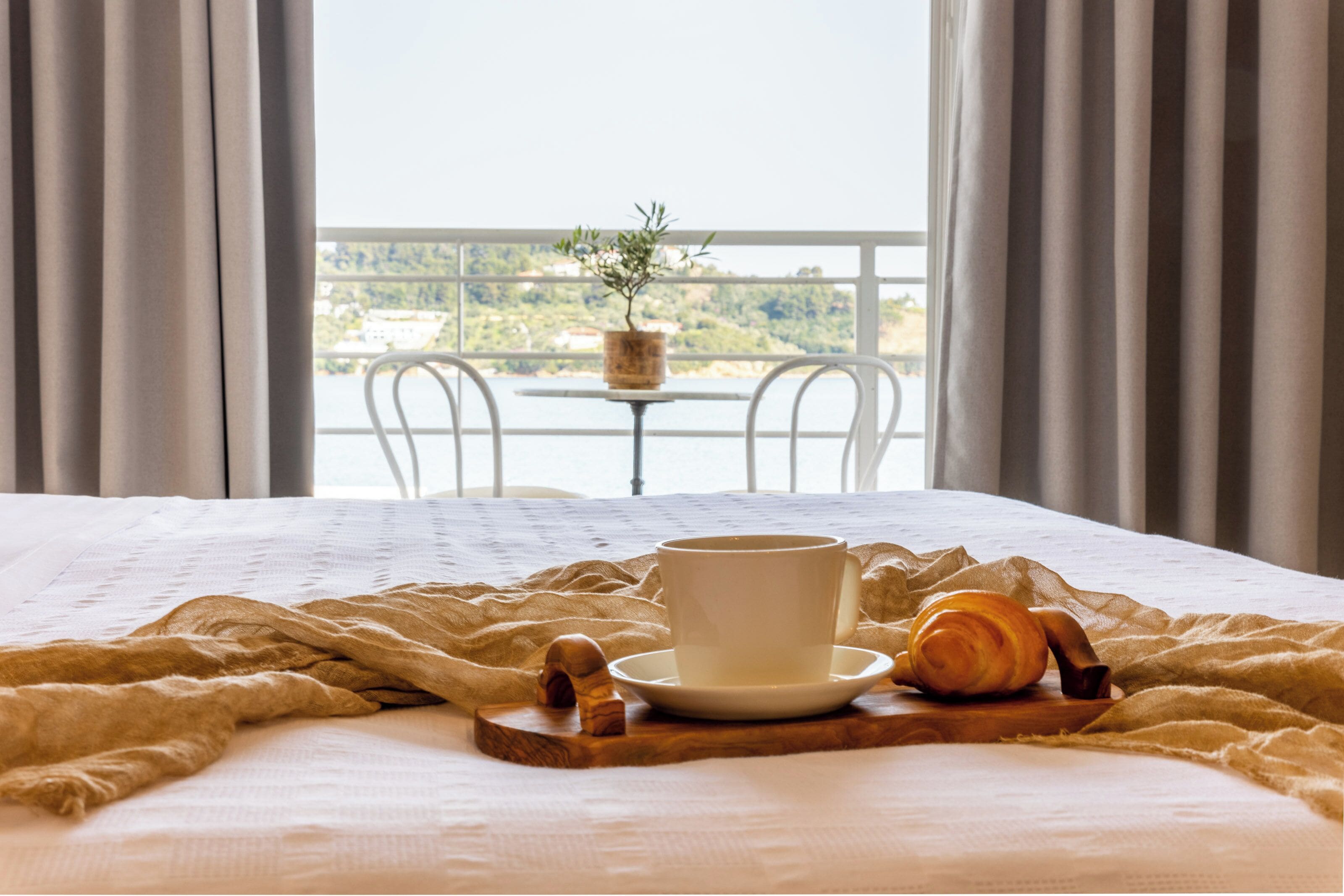 Una cama con edredón blanco, una taza de café y un croissant sobre una bandeja de madera.