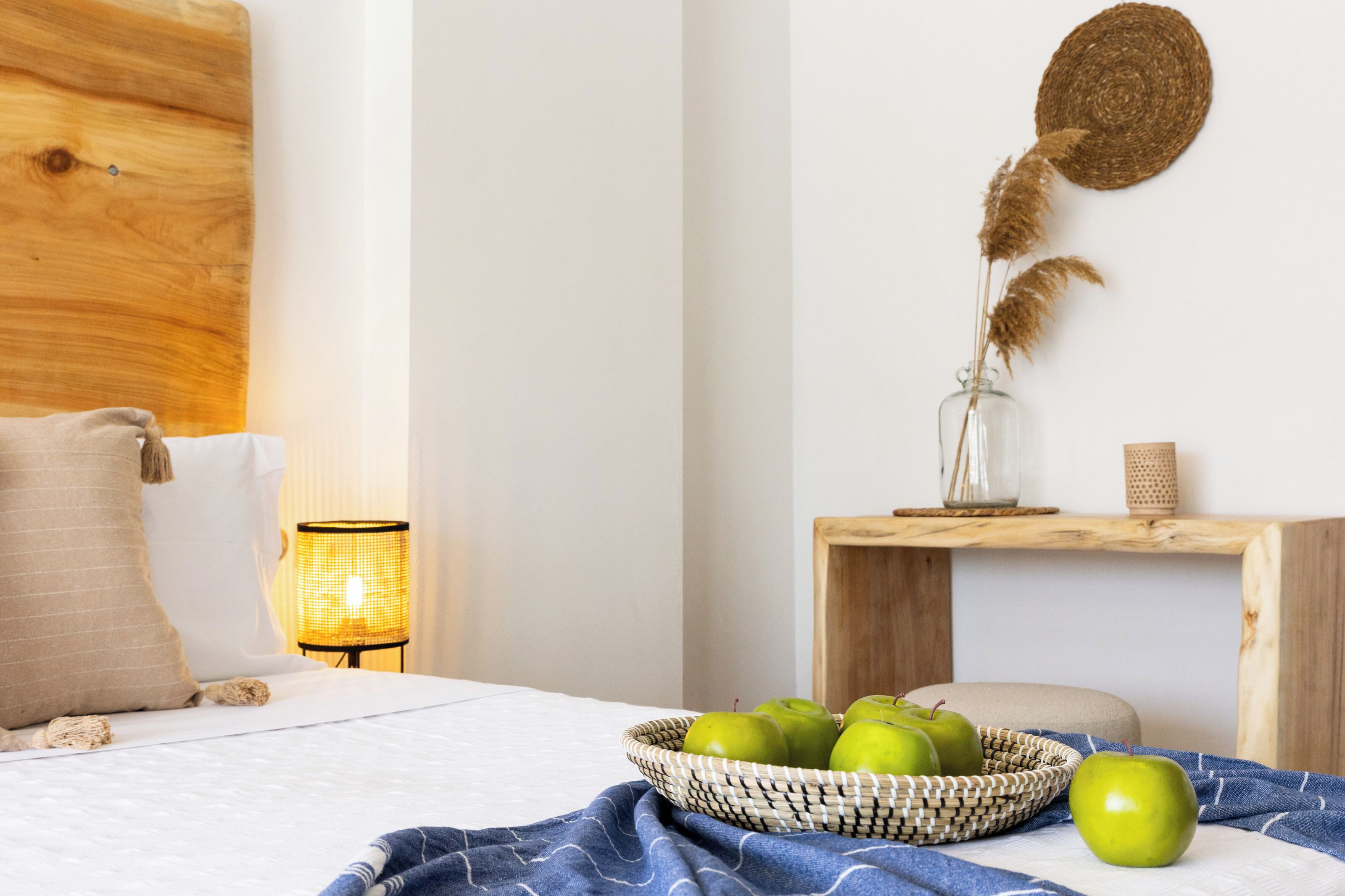 Een opgemaakt bed met een blauwe deken, een rieten mand met groene appels, een bedlampje en een houten nachtkastje met een gevlochten mand en decoratieve objecten.