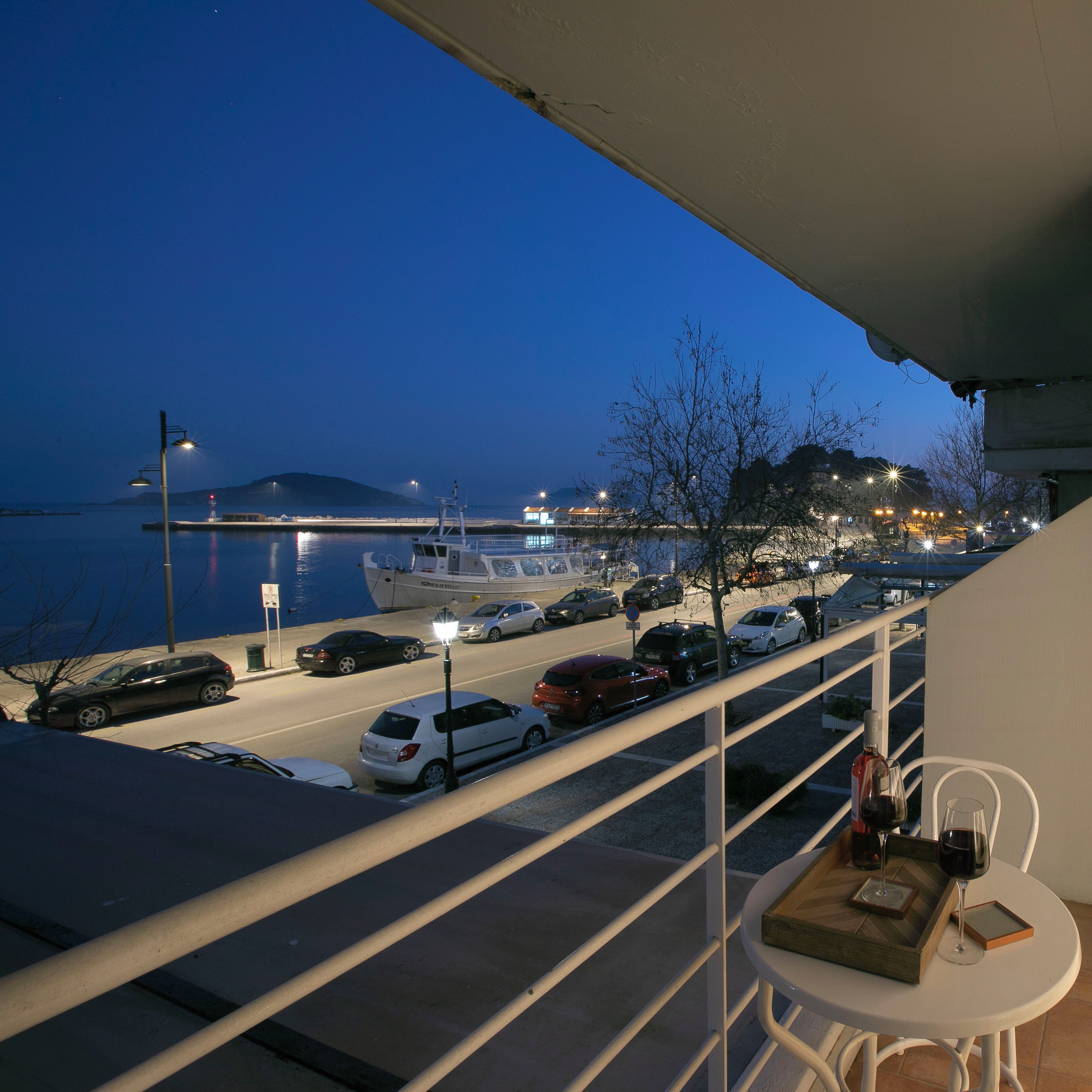A balcony with a view of a waterfront, cars parked along the street, and a table with a wine bottle and glass.