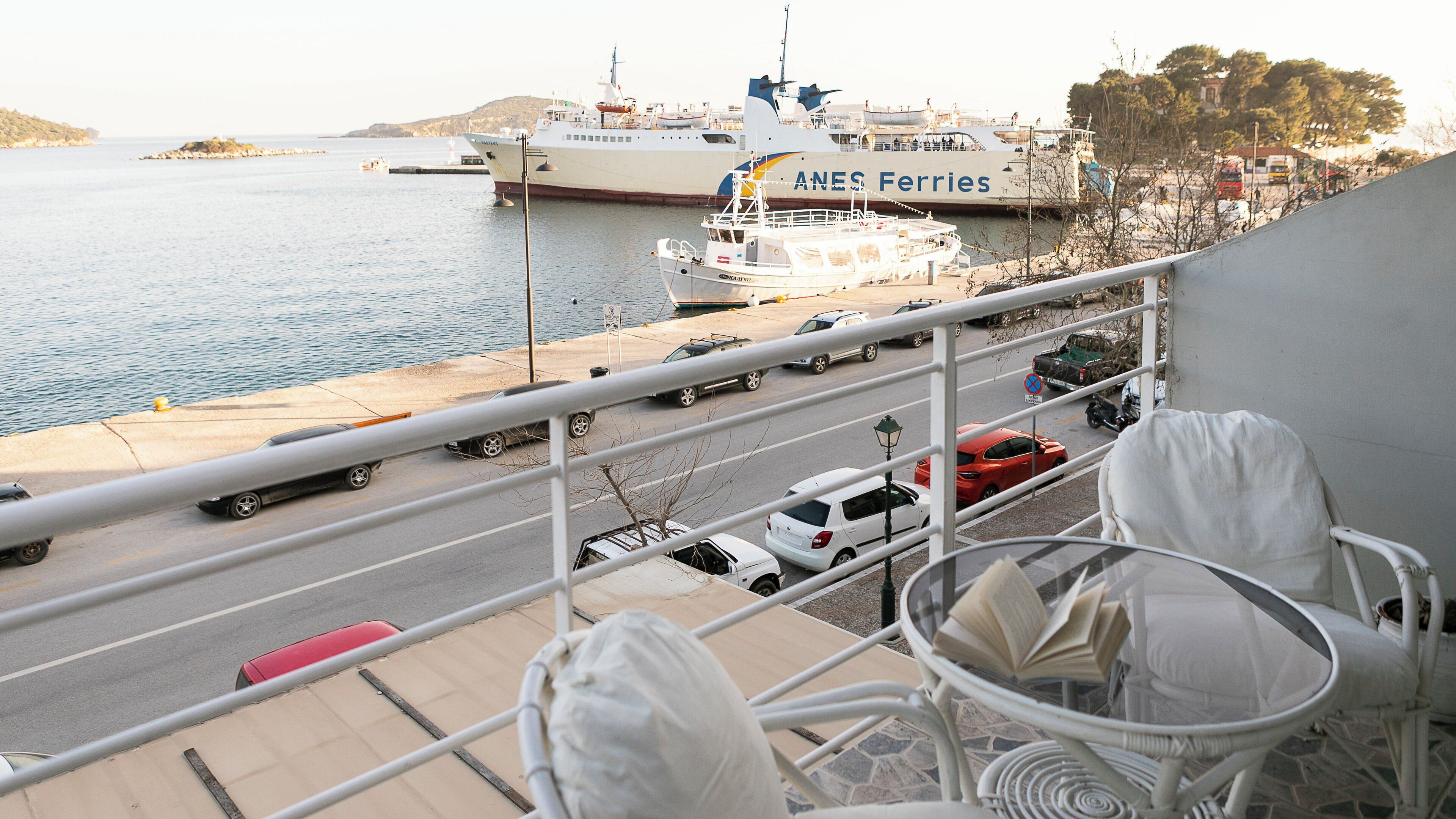 A balcony with a view of a ferry dock, a road with parked cars, and a white chair with a book.