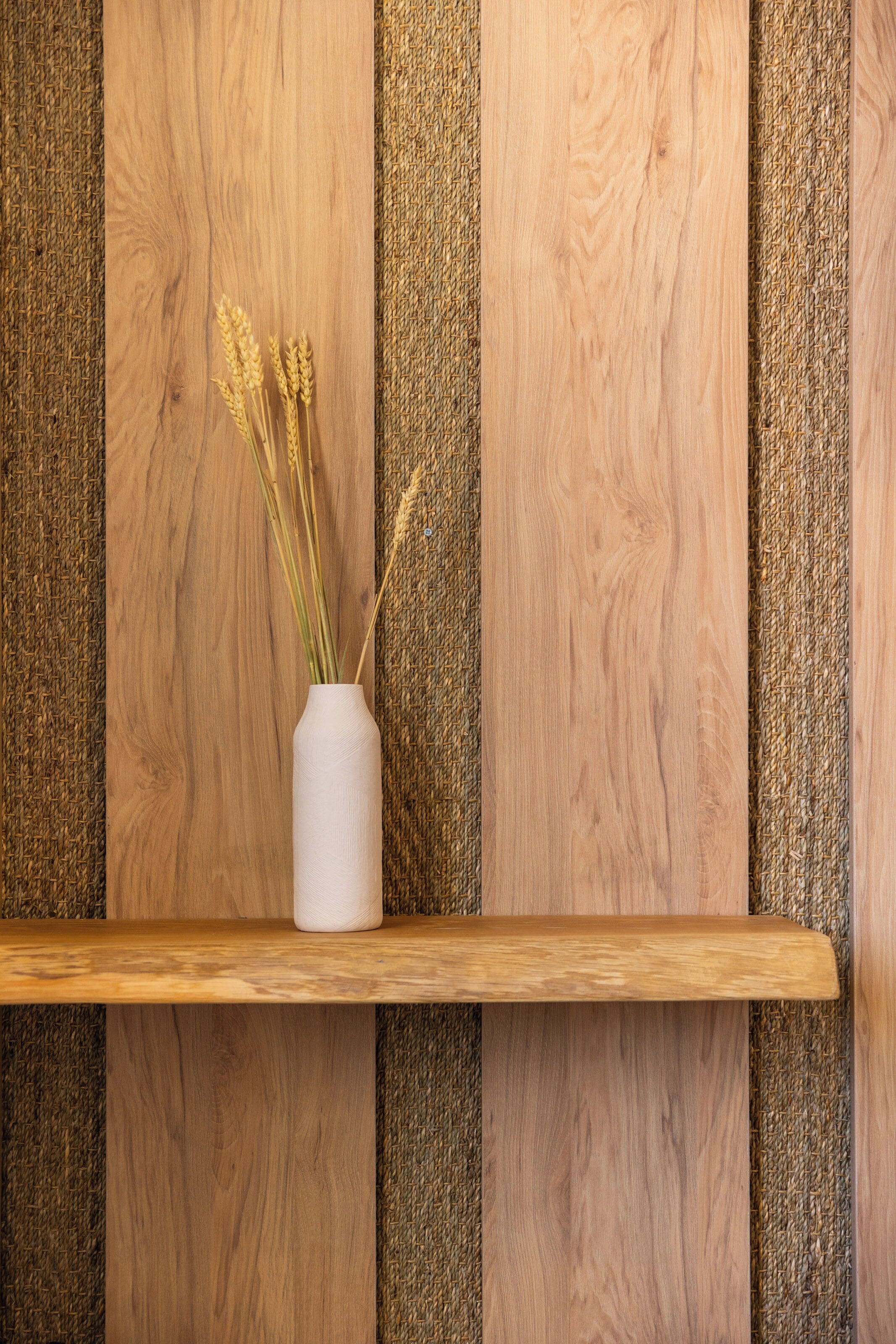Un jarrón blanco con plantas secas sobre un estante de madera frente a una pared con textura.