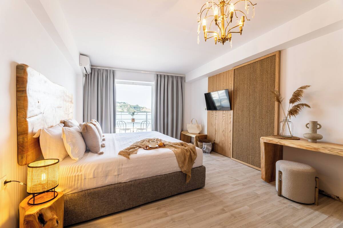 A modern bedroom with a large bed, a desk, a TV, and a view of the outdoors.