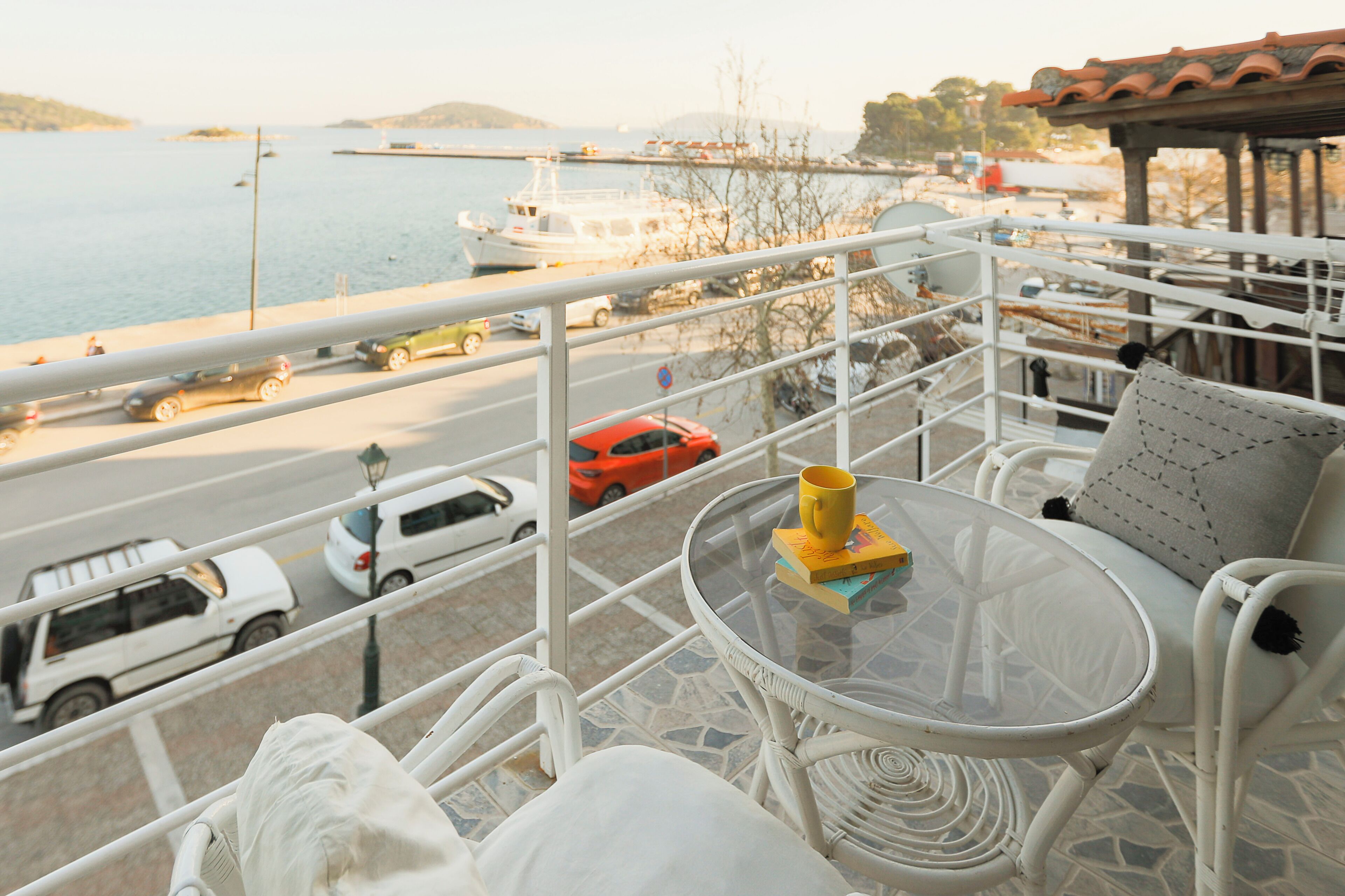 Een balkon met een glazen tafel, witte rieten stoelen, een gele beker en uitzicht op een jachthaven met boten en auto's.