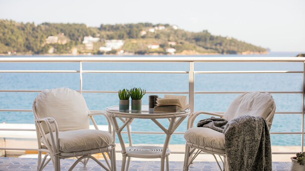 A balcony with two wicker chairs, a small round table with a book and two potted plants, and a view of the sea and a hillside. - Casa Blue Marine (Skiathos)