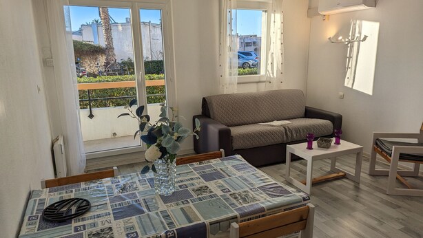 A living room with a sofa, a dining table with chairs, a small white table, and a balcony with a view of the outside.