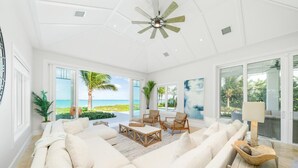 A spacious living room with a large white sofa, wooden coffee table, and armchairs. It features large windows with a view of the ocean and palm trees. - Seas The Day (Georgetown)