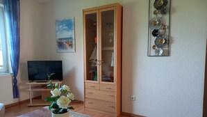 A living room with a wooden cabinet, a television, a table with a vase of flowers, and a wall with decorative items. - Vacation home Gerken - Apartment on the first floor (Drochtersen)