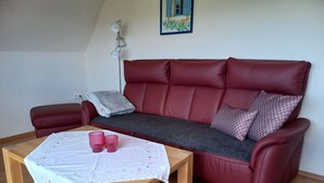 A red leather sofa with a grey throw blanket and patterned pillows, a wooden coffee table with a white tablecloth and pink glasses, and a painting on the wall. - Vacation home Gerken - Apartment in the attic (Drochtersen)