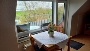 A dining area with a table set for two, a potted plant, and a view of a balcony with a railing and a body of water. - Vacation home Gerken - Apartment in the attic (Drochtersen)