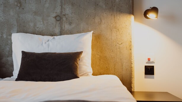 Una cama con almohadas blancas y oscuras, una mesita de noche y una lámpara fijada a la pared.