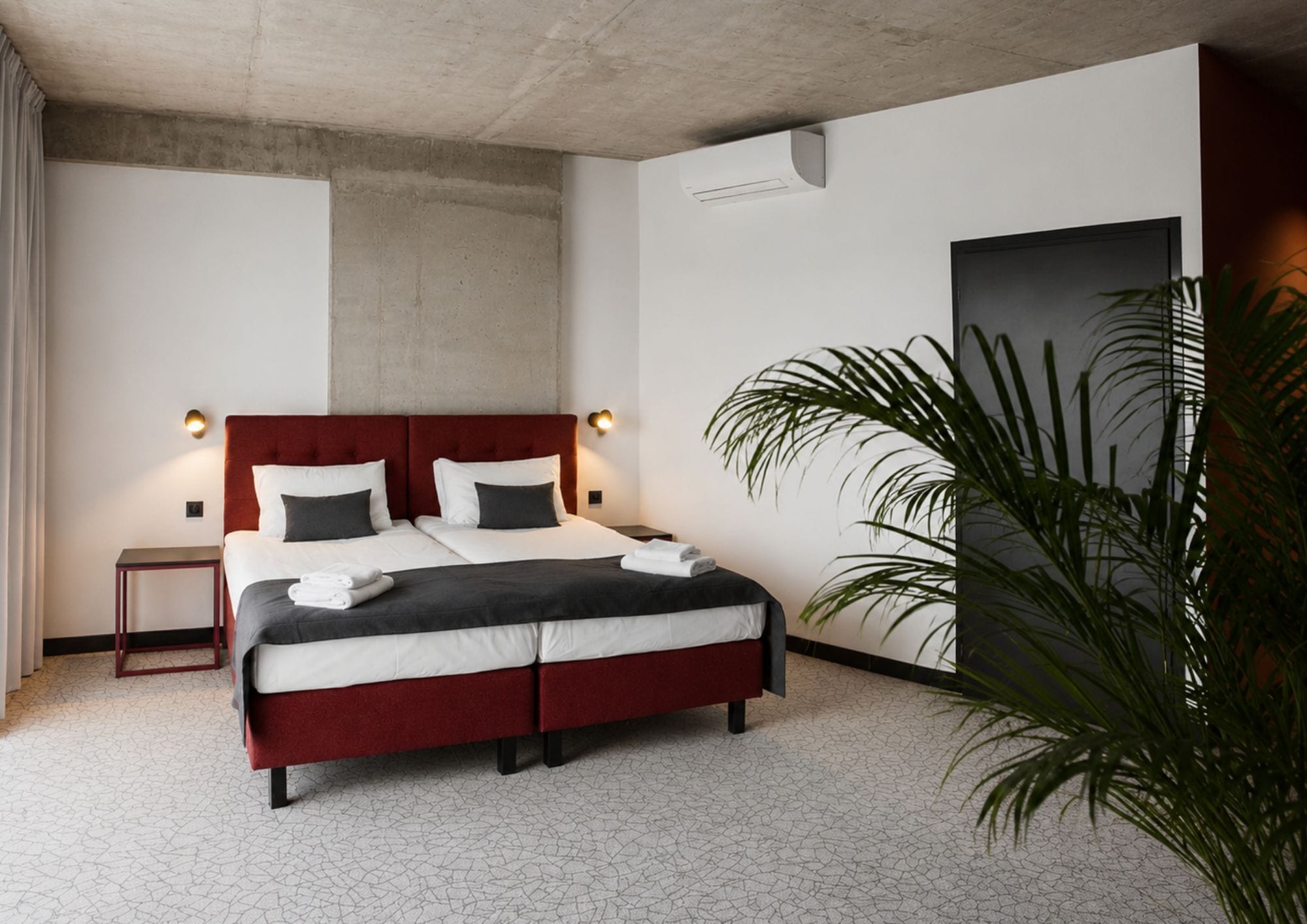 A modern hotel room with a large bed, two bedside tables, a small nightstand, a potted plant, and a large window.