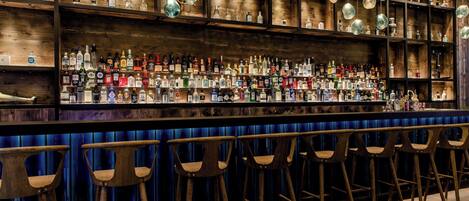 Uma área de bar com prateleiras de madeira repletas de garrafas diversas, luminárias pendentes esféricas e banquetas de bar de madeira.