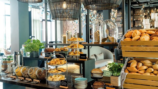 Een bakkerijbalie met verschillende gebakjes, waaronder croissants en brood, en een koffiehoek met een koffieapparaat en potten koffie.