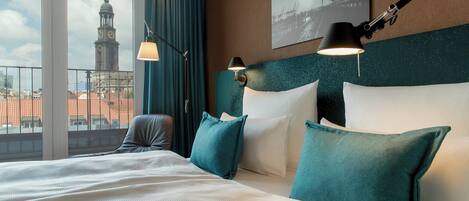 Habitación de hotel con una cama grande, almohadas color turquesa y vista a un paisaje urbano desde un balcón.