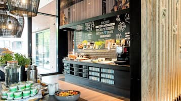 A breakfast bar with a variety of food items and beverages, a coffee machine, and a wall with multilingual greetings.