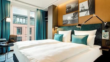 Ein Hotelzimmer mit einem großen Bett, einem Schreibtisch und einem Sessel. Zwei Fenster mit Vorhängen und Blick auf Gebäude.