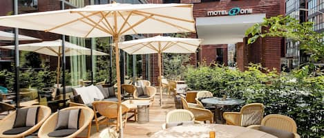 An outdoor seating area with wicker furniture and umbrellas, a brick building in the background, and a sign for 'HOTEL ONE'.