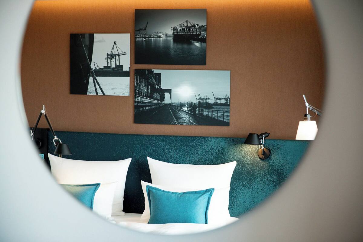 Une chambre d’hôtel avec un lit, des oreillers, une lampe fixée au mur et trois photographies en noir et blanc représentant des scènes industrielles.