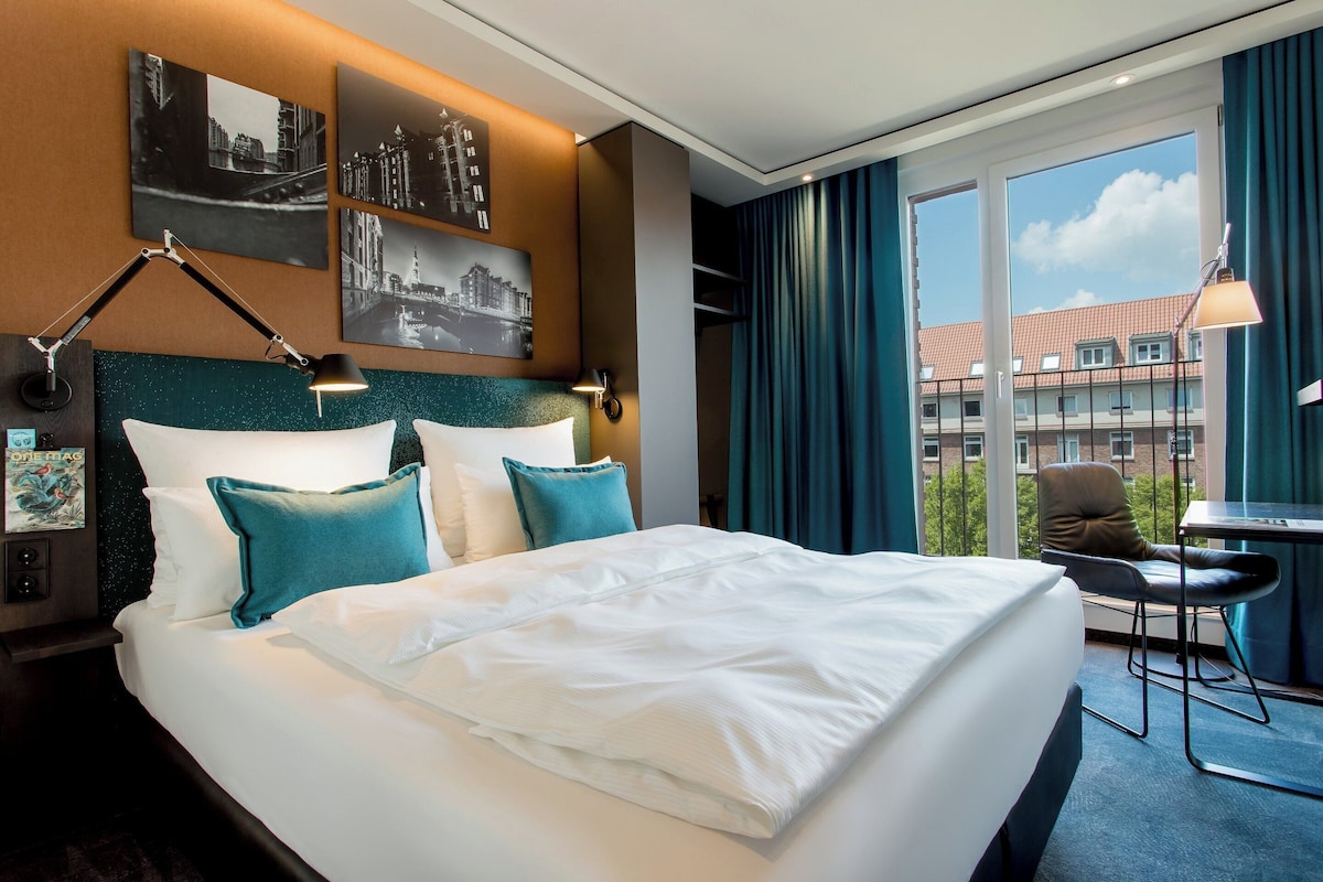Habitación de hotel con una cama grande, dos almohadas, una mesita de noche, un escritorio, una silla y una ventana con cortinas.