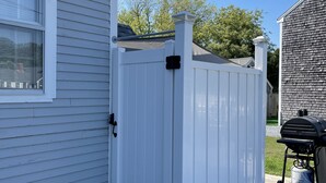 A white wooden fence with black corner brackets, a grill cart with propane tank, and a house with white siding. - Lower County Bunk House (Dennis)